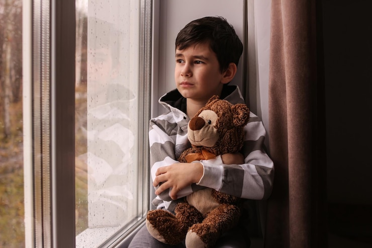 Foto de jongen kijkt droomend uit het raam en knuffelt een teddybeer.