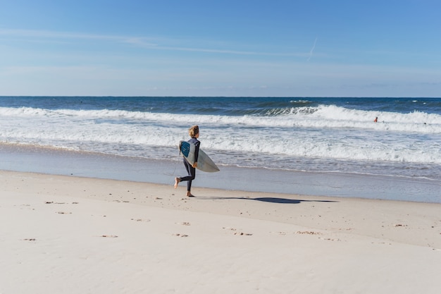 de jongen knielt op het strand voordat hij gaat surfen en rent het water in met een surfplank