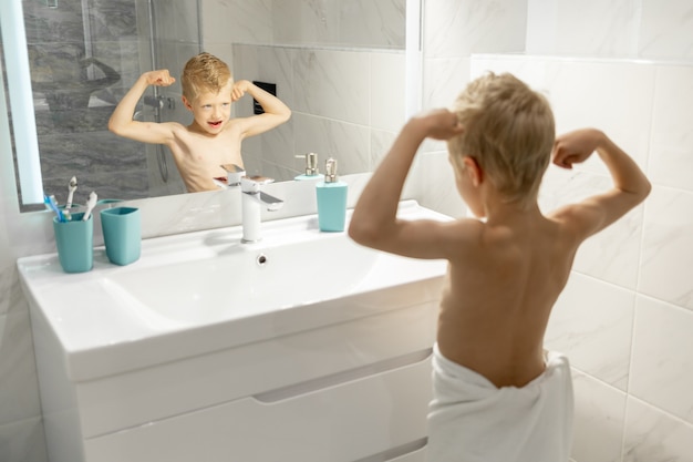 De jongen laat zijn biceps zien of trekt een gezicht voor de badkamerspiegel