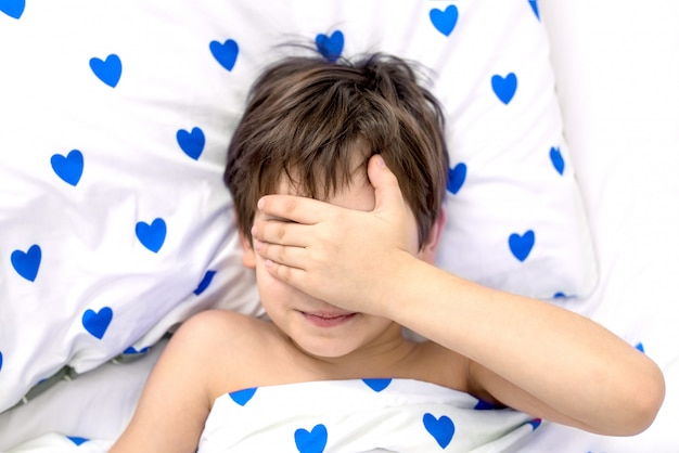 de jongen ligt in een bed met blauwe harten, zijn gezicht in zijn handen. emoties zonder een gezicht. witte kleur, bovenaanzicht