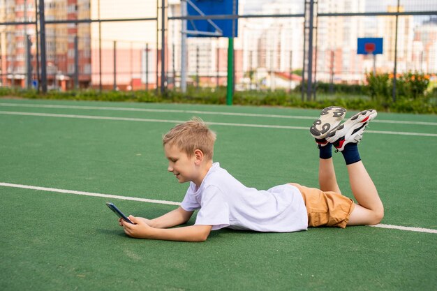 De jongen ligt op het groene gras om te voetballen en aan de telefoon te spelen