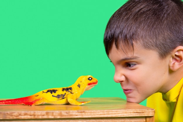 Foto de jongen met korte snit in geel shirt kijkt aandachtig naar een dinosaurusspeelgoed dat zijn kin op tafel legt