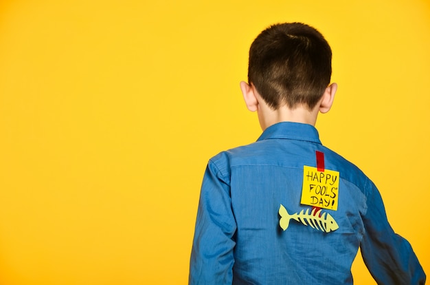 De jongen op een gele achtergrond in een blauw shirt met een vis gelijmd tape en een stuk papier op zijn rug