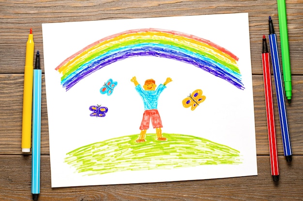 De jongen verheugt zich in de regenboog Kindertekening op wit papier Houten tafel met stiften