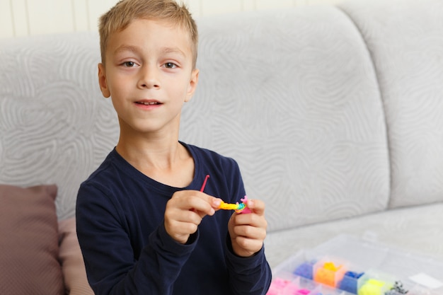 De jongen weeft armband van rubberringen thuis op de bank