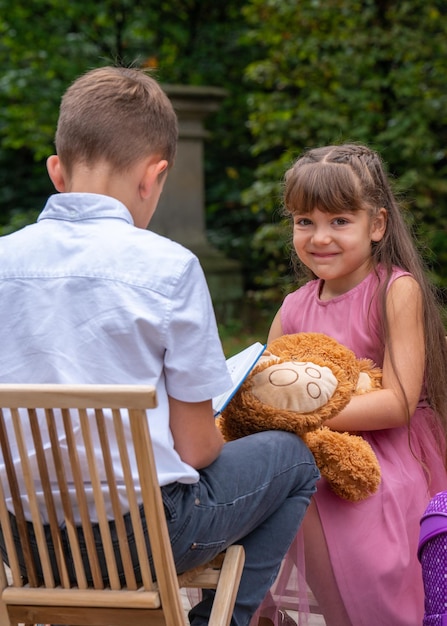 De jongen zit met zijn rug op een stoel en leest een boek. Het meisje gluurt erin en wil het zien