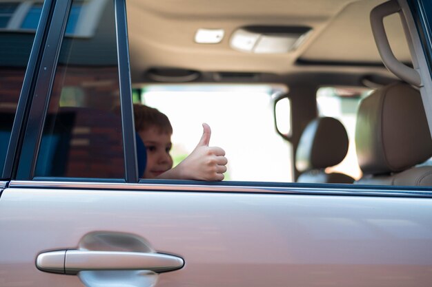 De jongen zit op de achterbank van een zilveren auto en laat een duim zien Kleuter is klaar voor een spannende reis