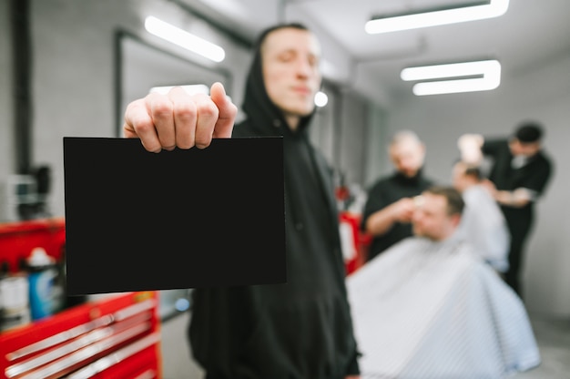 De kapper in zwarte hoodie houdt zwarte kaart op de achtergrond van de kapperswinkel en kappers die cliënten knippen. Man houdt een blanco kaart in zijn hand op een achtergrond van een mannelijke kapper.