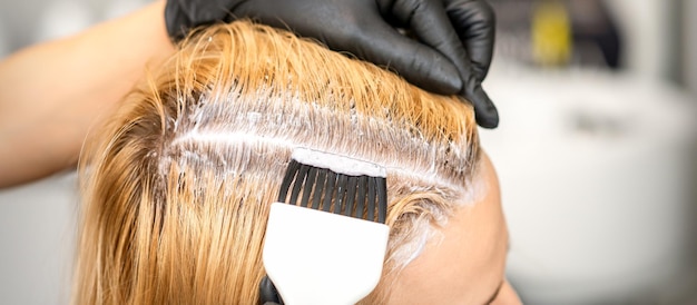 De kapper verft blonde haarwortels met een borstel voor een jonge vrouw in een kapsalon