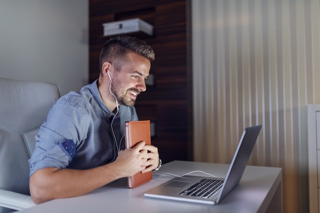 De Kaukasische gebaarde agenda van de werknemersholding in handen, hebbend videogesprek over laptop en zittend in bureau laat in de nacht.