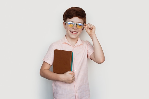 De Kaukasische gemberjongen met bril glimlacht bij camera die enkele boeken op een witte studiomuur in vrijetijdskleding houdt