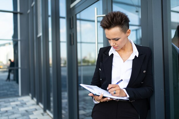 De Kaukasische vrouw in bureaukleren schrijft aan notitieboekje en wacht collega dichtbij het bureaugebouw