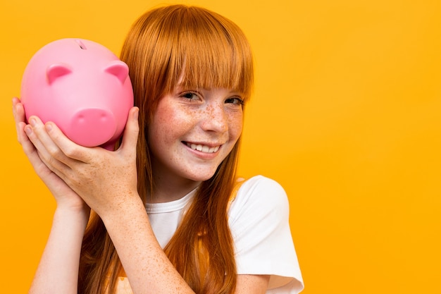 De Kaukasische vrouw met rood haar houdt roze die varkensspaarpot op geel wordt geïsoleerd