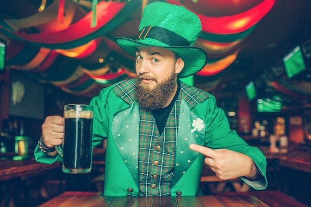 De kerel in groen kostuum zit bij lijst in bar en het stellen. hij houdt een mok donker bier vast en kijkt. hij is zelfverzekerd.