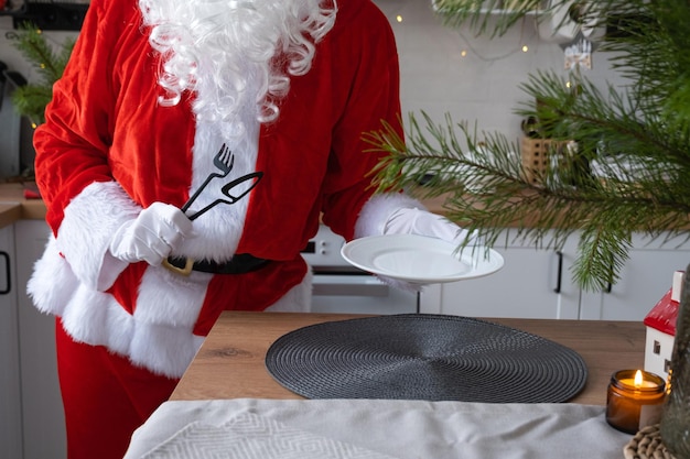 De kerstman dekt de tafel thuis in de witte keuken zet een bord neer zet bestek vork en mes Voorbereiding op de viering van Kerstmis de tafel dekken voor gasten