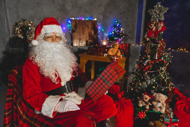 De kerstman houdt een geschenkdoos thuis Zit op een stoel denk na over een plan voor een verzonden geschenkdoos om kinderen te geven