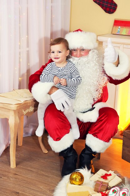 De kerstman houdt een klein schattig meisje vast bij de open haard en de kerstboom thuis Christmas