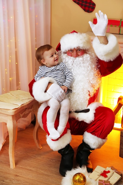 De kerstman houdt een klein schattig meisje vast bij de open haard en de kerstboom thuis