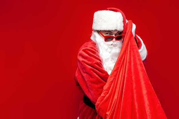 De Kerstman in een feestelijke bril en hoed haalt cadeautjes uit de tas op rode achtergrond