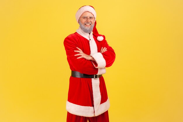 De kerstman kijkt direct naar de camera, houdt de handen gevouwen, blije zelfverzekerde uitdrukking.