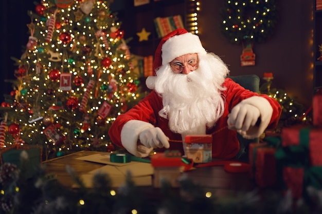 De Kerstman of Sinterklaas maakt cadeaus terwijl hij thuis bij de kerstboom aan tafel zit