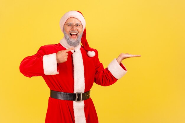 De kerstman presenteert kopieerruimte op zijn handpalm, kijkend naar de camera met open mond.