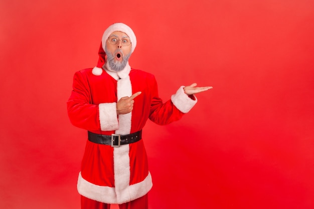 De kerstman staat met open mond wijzende wijsvinger opzij op kopieerruimte.