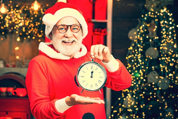 De kerstman trekt een grappig gezicht en houdt de klok vast die vijf minuten voor middernacht laat zien. de kerstman houdt een alarm vast