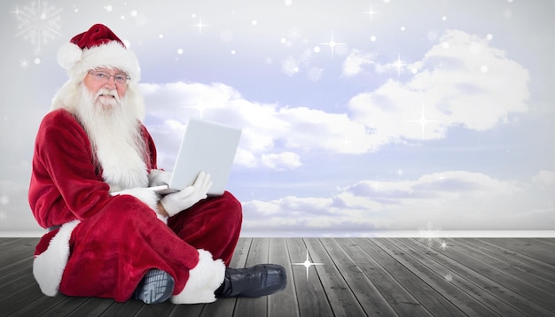 De kerstman zit en gebruikt laptop tegen wolken in een ruimte