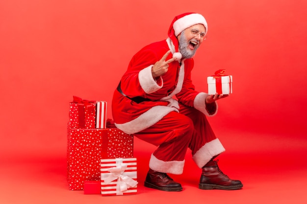 De kerstman zit op een grote doos met kerstcadeau, houdt een cadeau vast en toont een v-teken aan de camera.