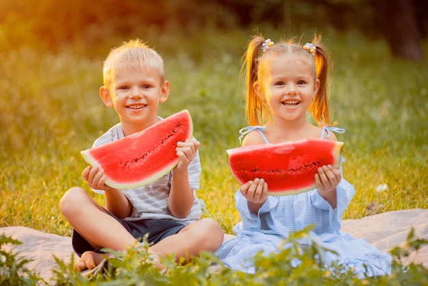 De kinderen op het gazon met plakken van watermeloen in hun dienen de stralen van zonsondergang in