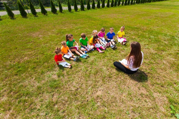 De kinderen zitten in een rij op het gras tegenover de juf en kijken in hun notitieboekjes