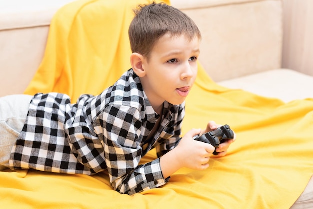 De kindjongen in geruit overhemd liggend op de bank met zwarte joystick in zijn handen die het spel speelt