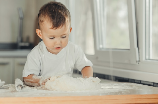 De kleine jongen in de keuken kneedt zelf in een wit t-shirt hard het deeg