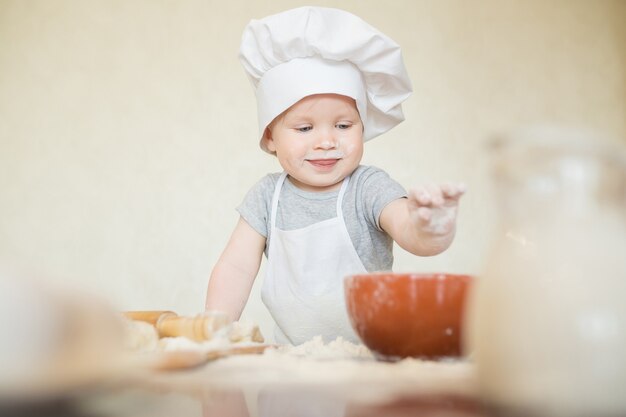 De kleine jongen in een pak van de kok beeldhouwt deeg. Baby maakt diner in kokskostuum