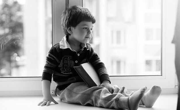 De kleine jongen leest een boek. Het kind zit bij het raam en bereidt zich voor op lessen. Jongen met een boek in zijn handen zit op de vensterbank.