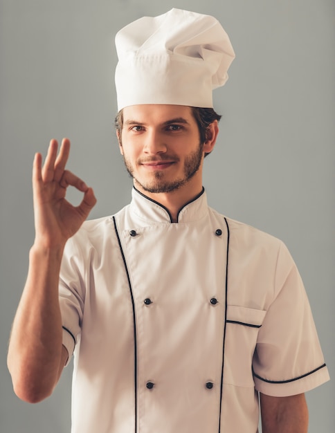 De knappe jonge kok in uniform toont ok teken.