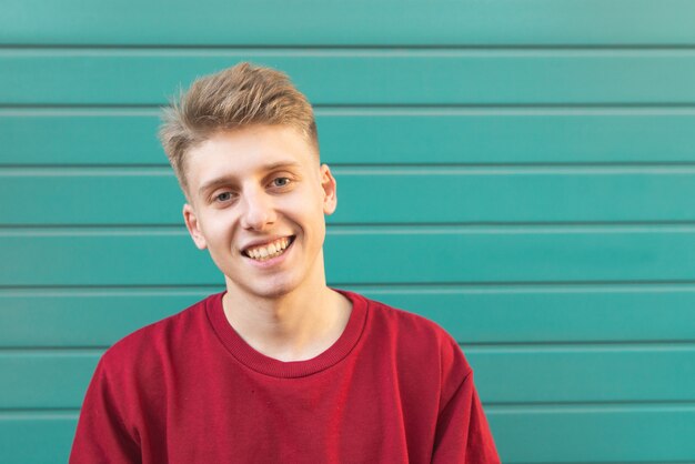 Foto de knappe jonge mens in een rood sweatshirt op turkooise muur ziet eruit en glimlacht