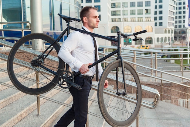 De knappe jonge zakenman in een wit overhemd en een avondkleding draagt zijn fiets