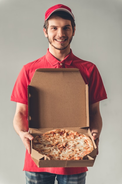 De knappe leveringsarbeider in rood uniform houdt een pizza.