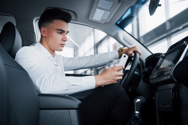 De knappe man is een koper die in een auto zit met een telefoon in een nieuwe auto bij een dealercentrum.