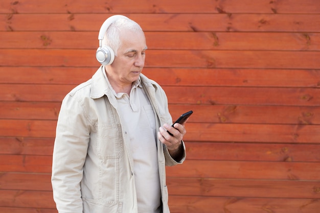 De knappe oude mens gekleed in slimme toevallige stijl gebruikt een smartphone en draagt hoofdtelefoons