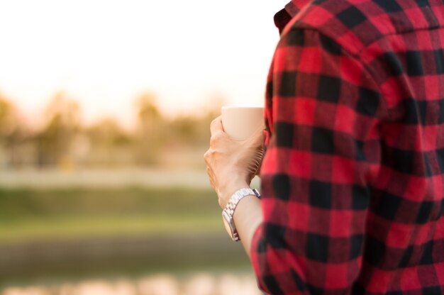 De koffiekop van de mensengreep in hand bevindt zich dichtbij het meer met ochtendlicht.