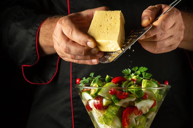 De kok voegt kaas toe aan een vitaminesalade voor het avondeten Het concept van het bereiden van een vegetarisch gerecht met de handen van een chef-kok Groentengerecht in een kom