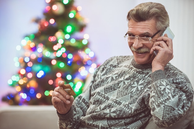 De lachende oude man met een creditcardtelefoon bij de kerstboom