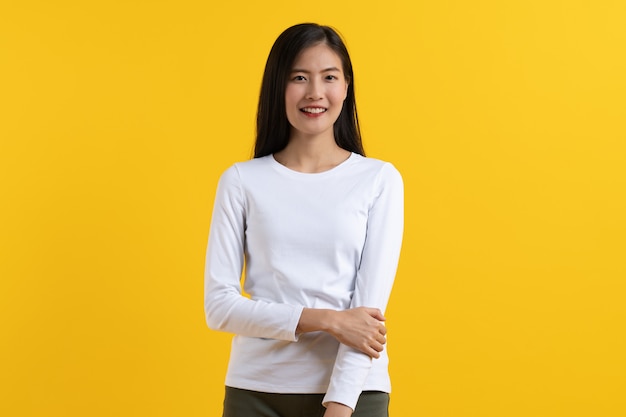 De leuke Aziatische jonge vrouw in wit toevallig overhemd stelt stellend vertrouwen gevouwen wapens die op gele achtergrond in studio worden geïsoleerd.