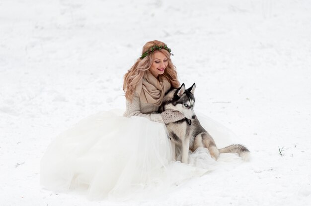 De leuke bruid met kroon speelt met siberische schor op achtergrond van witte sneeuw. winter bruiloft.