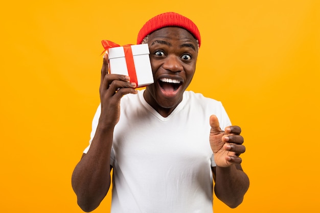 De leuke verraste zwarte Afrikaanse mens met een glimlach in een wit t-shirt houdt een doos een gift met een rood lint voor een gele verjaardag