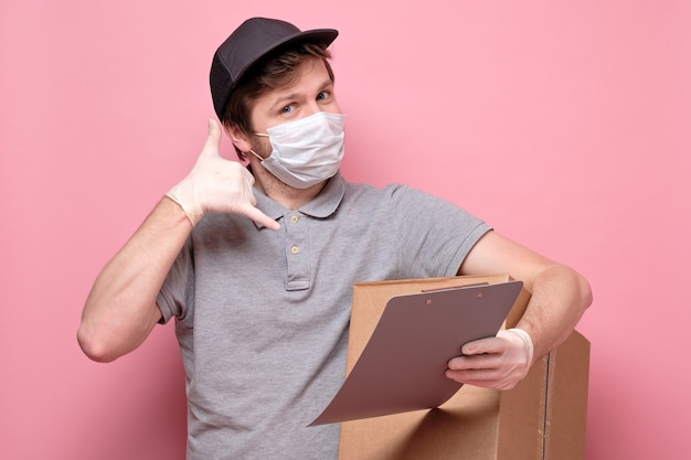 De leveringsmens in medisch masker die een cardbox houden die tonen telefoneert me gebaar