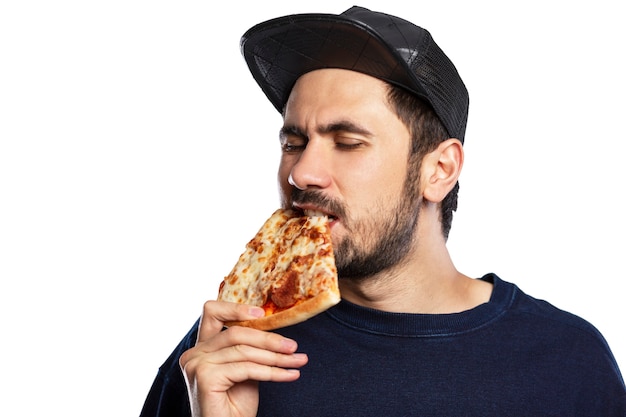 De man eet een stuk pizza. Jonge brunette met een baard en in een pet. Smakelijk tussendoortje. Detailopname. Geïsoleerd op een witte achtergrond. Ruimte voor tekst.